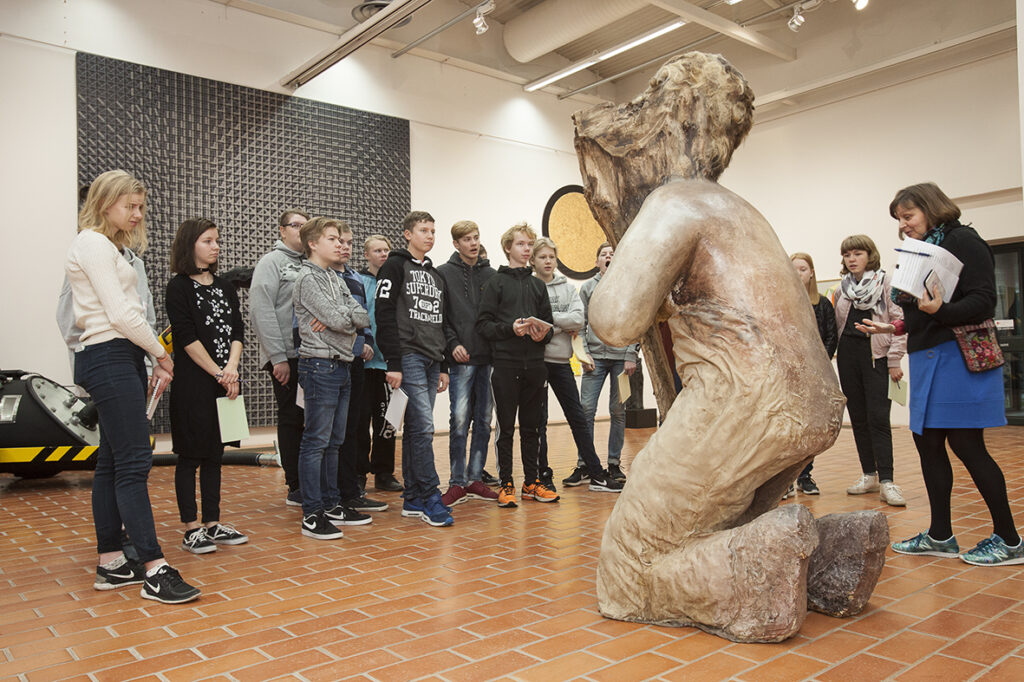 A group of young people are gathered around a large sculpture in an exhibition at Pori Art Museum. The 2-meter-tall sculpture is made out of wood, polyurethane and fabric and it resembles a kneeling human figure with its side facing the photographer.