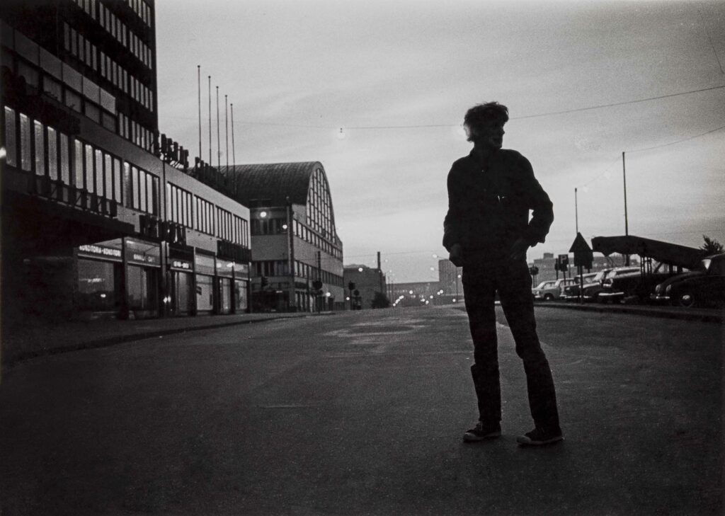 Jan Eeralan mustavalkoinen valokuva vuodelta 1966. Mies seisoo kaupungin kadulla yöllä tai illan hämärässä. Henkilö on kuvassa etualalla ja katsoo sivulle. Henkilön tumma profiili erottuu kaupunkisiluettia vasten.