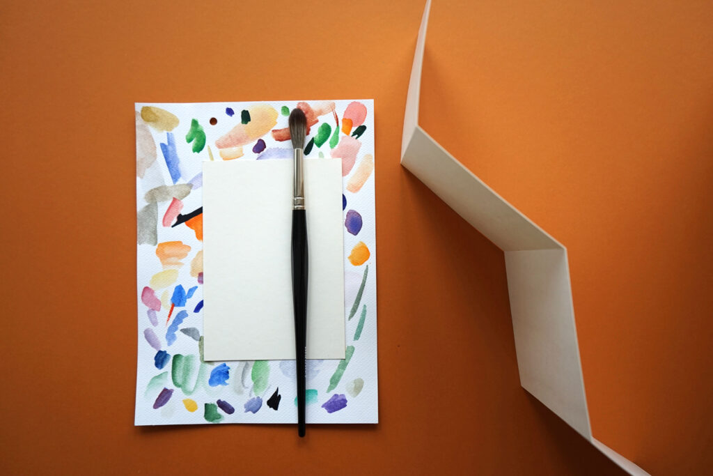 Vesivärimaalaus, valkoinen paperi ja sivellin ylhäältäpäin kuvattuna. Kuvan tausta on oranssinruskea.
