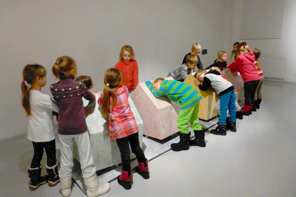 Vaaleassa näyttelytilassa on viisitoista päiväkotilasta. Lapset halaavat, nojaavat, kumartuvat kohti, liikuttelevat tai muuten koskevat heidän keskellään olevaa Taajama-teosta. Teos on lapsia matalampi. Teoksen neljä osaa ovat talon mallisia ja niiden alla on suorakaiteen muotoinen levy.