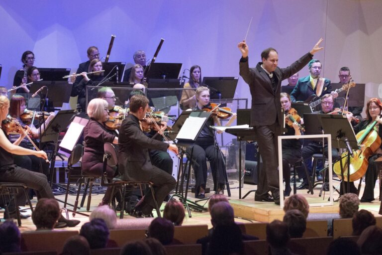 Pori Sinfonietta konsertoi Promenadisalin lavalla kapellimestarinaan Tibor Boganyi, joka on kääntynyt kohti yleisöä hymyillen kädet ylhäällä. Salin valaistus violetti.