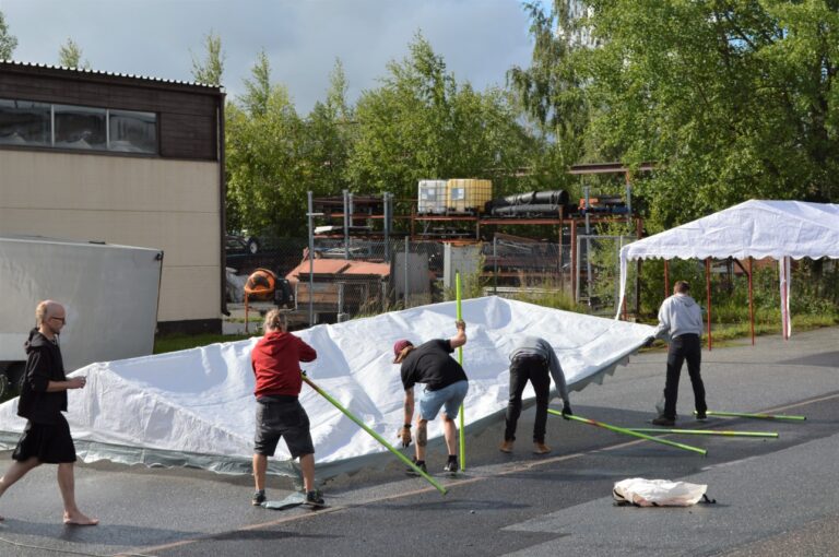 Neljä ihmistä pystyttää telttakatosta asvaltille aurinkoisena kesäpäivänä