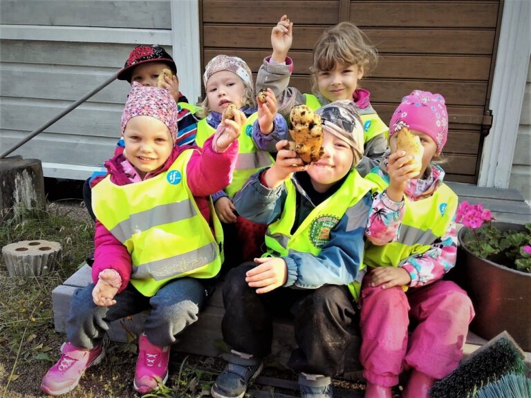Kuusi hymyilevää lasta istuu ulkona portailla ja nostavat ilmaan perunoita