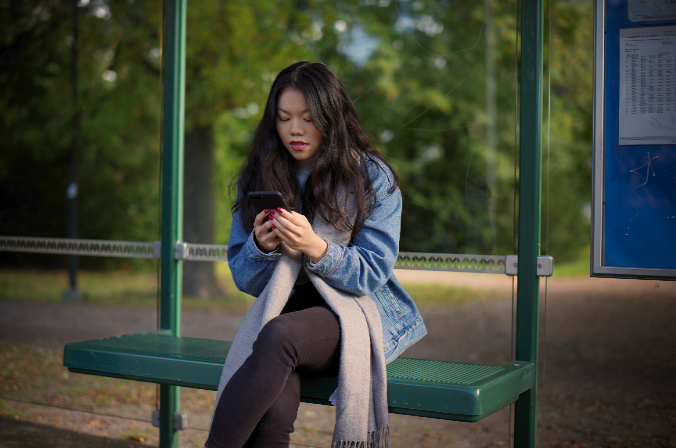 Nainen istuu bussipysäkillä kännykkä kädessä