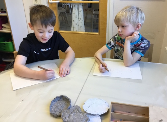Kaksi lasta pöydän äärellä sisätiloissa piirtää kynällä paperille päiväkodin tiloissa.