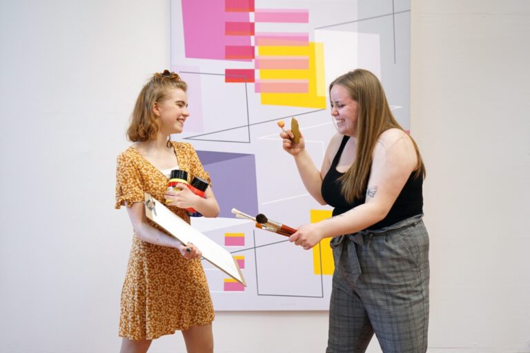 Kaksi nuorta naista katsovat toisiaan ja hymyilevät abstraktin maalauksen edessä, sisätila, naisilla käsissään taidemaalaustarvikkeita.