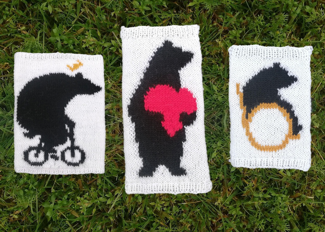 Ruohikolla kolme valkoista käsityötä, joissa jokaisessa yksi musta karhu. Yksi karhu kelaa pyörätuolissa, yksi pitää sylissä punaista sydäntä, yksi ajaa polkupyörää. 