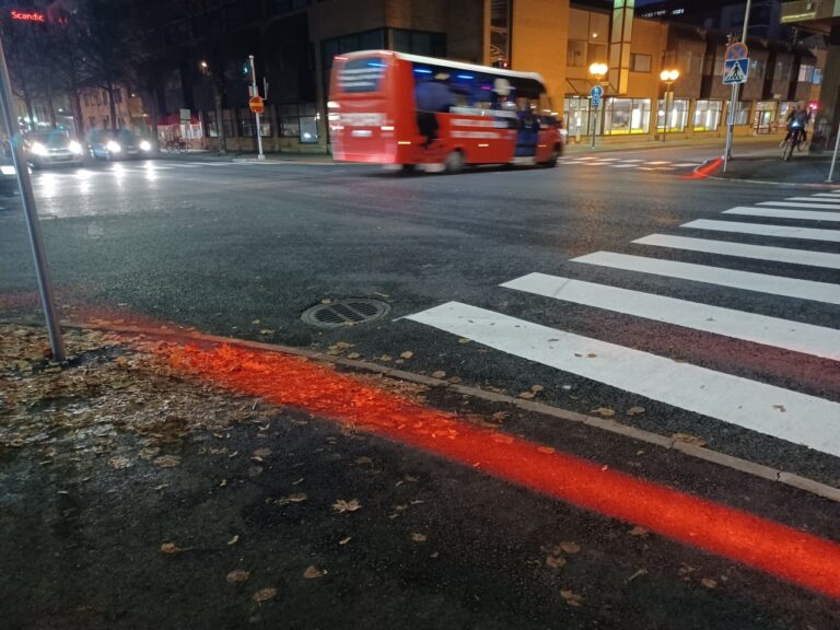 Risteysalue pimeänä syysiltana. Risteyksessä punainen pieni citylinja-bussi. Punainen valo heijastaa maahan suojatien molemmissa päissä.