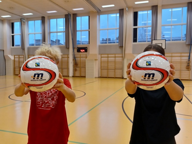 Kaksi lasta pitää koulun liikuntasalissa käsissään reilun kaupan urheilupalloja. 