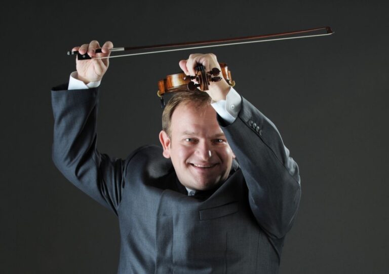 Kapellimestari-viulisti Michael Maciaszczyk pitää viulua päänsä päällä hymyillen.