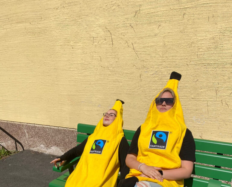 Kaksi keltaiseen banaanipukuun pukeutunutta ihmistä nauttii auringosta vihreällä penkillä.