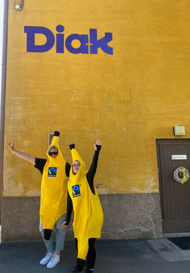 Kaksi keltaiseen banaanipukuun pukeutunutta ihmistä poseeraa Diak-kyltin alla. 