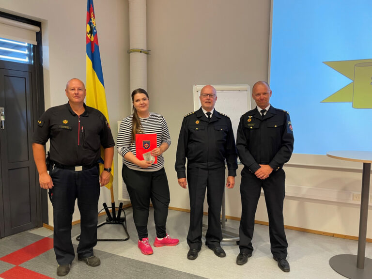 Tiina Koivumäki ja pelastuslaitoksen edustajat ryhmäkuvassa Satakunnan pelastuslaitoksen lipun edessä.