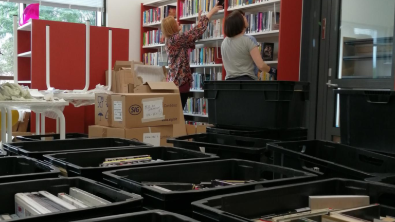 Kaksi kirjaston työntekijää tyhjentämässä kirjoja muuttolaatikoista kirjahyllyihin