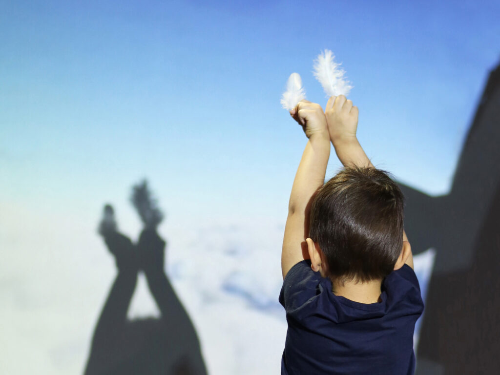 A child makes a shadow to the wall by lifting up his hands with feathers in them.