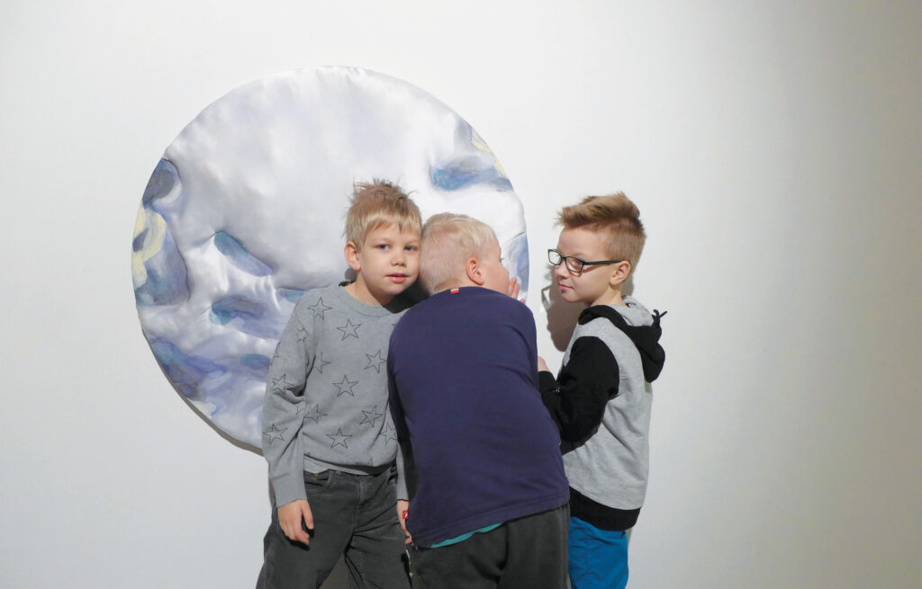 Kolme nuorta poikaa kuuntelee ja koskettaa seinällä olevaa taideteosta.