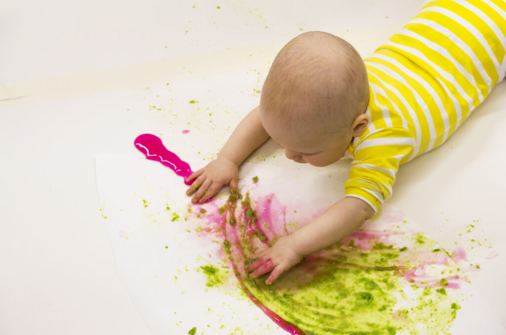 A baby lies on the ground on its tummy and paints with their hands.