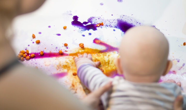 Vauva maalaa käsillään aikuisen tukiessa lapsen asentoa. Värikkäät maalausaineet sisältävät muun muassa tyrnimarjoja.