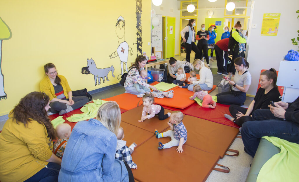 Lots of families with babies are sitting on the floor of the child health clinic.