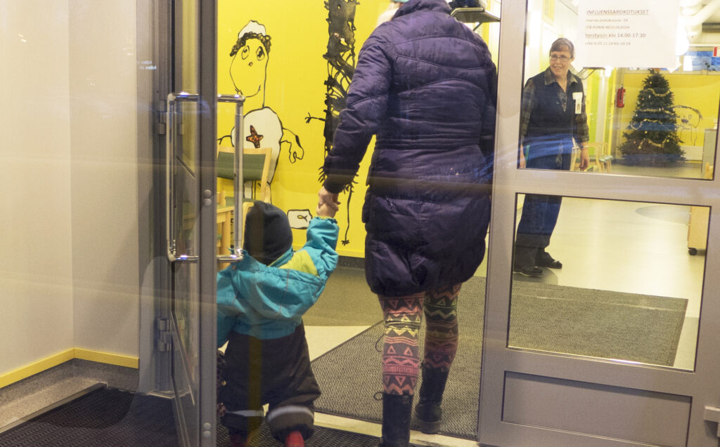 Lapsi ja aikuinen kävelevät käsi kädessä neuvolan ulko-ovesta sisään. Neuvolan seinillä on taidetta.
