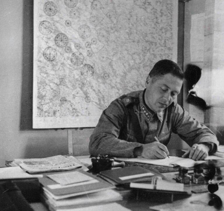 Asepukuinen mies istuu työpöydän äärellä takanaan Porin kaupungin kartta. Työpöydällä on puhelin, asiapapereita ja muistiinpanovälineitä..