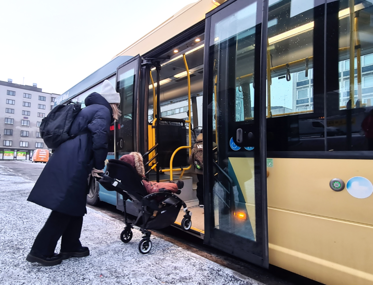 Mustaan talvitakkiin ja vaaleaan pipoon pukeutunut nainen työntää lastenrattaita sisään linja-auton keskiovista talvella.