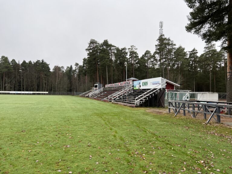 Nurmikenttä jonka taustalla metsäisiä puita, katsomo jossa kyltti me ollaan musa nyt ja aina