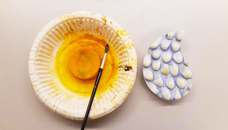 Paperilautasella on keltainen vesivärinappi ja pensseli, pöydällä on myös pisaranmuotoinen kipsiveistos, jota on maalattu sinertäväksi.