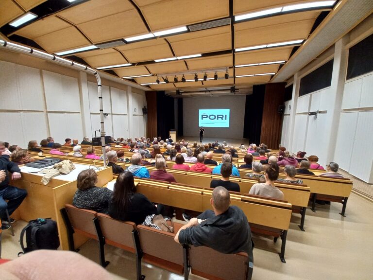 Kuva auditoriosta, jossa ihmisiä istumassa ja edessä näkyy iso kalvo jossa Porin logo.
