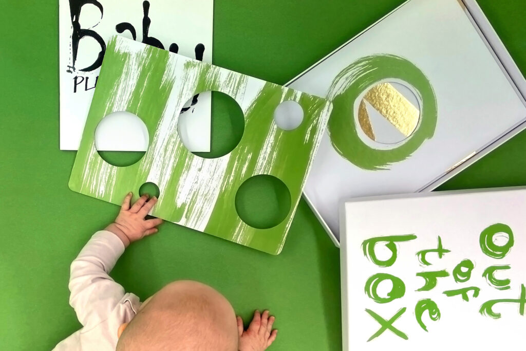vihreä tausta jossa valkovihreä pahvilaatikko ja etualalla vauva leikkimässä sillä