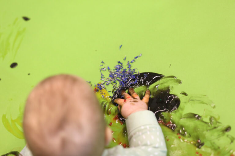vihreä tausta, edessä vauvan pää ja käsi ja hän leikkii värimateriaaleilla