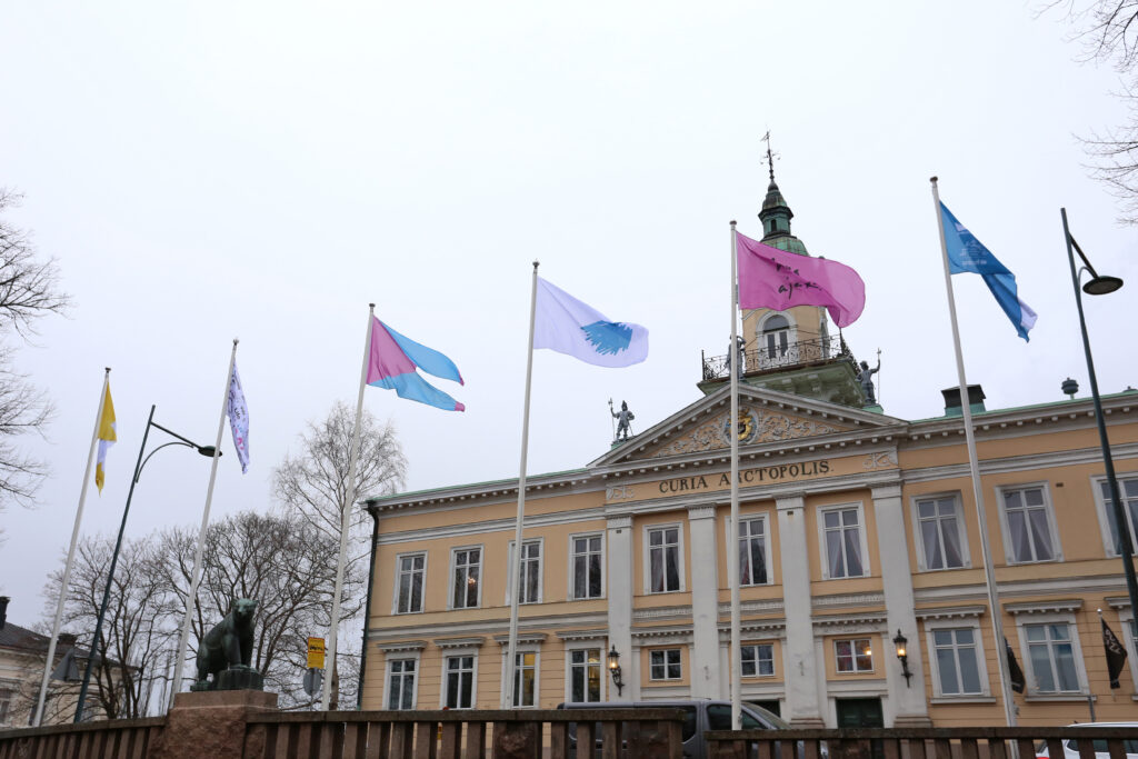 Keltaisen vanhan raatihuonerakennuksen edessä kuusi lipputankoa joissa erilaisia lippuja