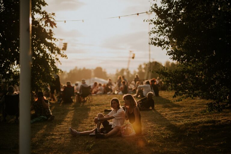 Kesäillan tunnelmaa festivaalialueelta. Ihmisiä istuskelemassa ruohikolla ja taustalla laskee aurinko.